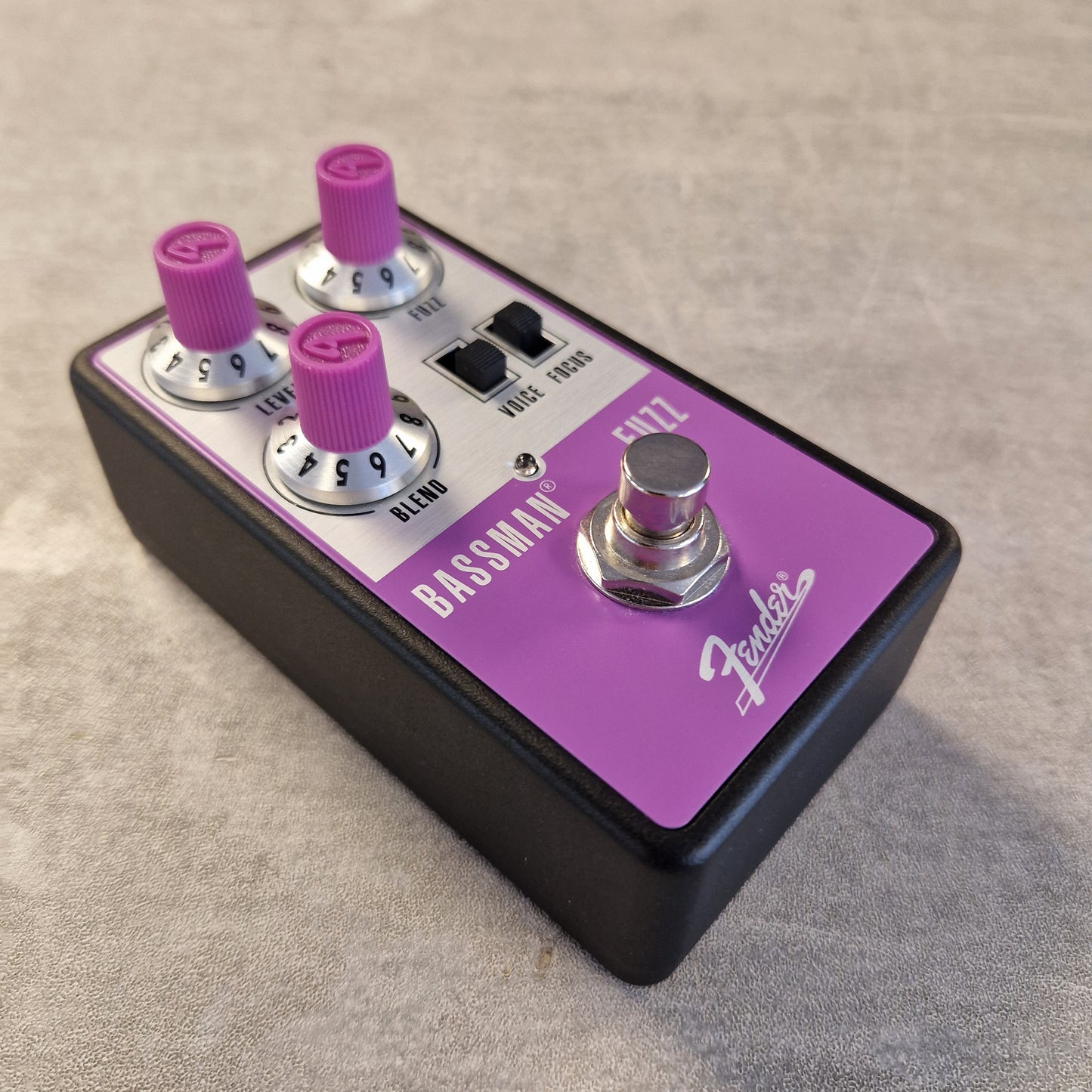 Pre-Owned Fender Bassman Fuzz Pedal Fender Bassman EZ72 bass guitar effects pedal with a black body, purple control panel, and pink knobs, shown on a textured gray surface.