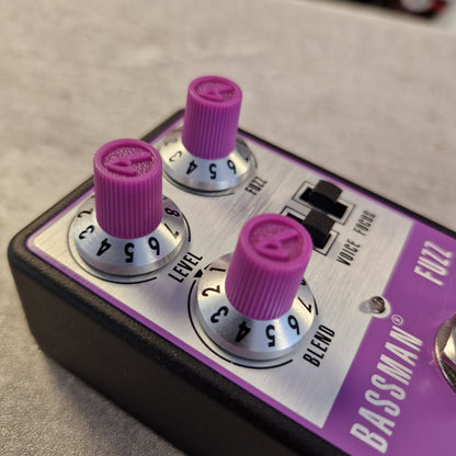 Pre-Owned Fender Bassman Fuzz Pedal Close-up of a Bassman Fuzz guitar effects pedal with three purple rotary knobs, black switches, a silver control panel, and a purple side plate on a dark base.