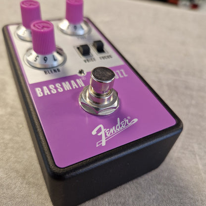 Pre-Owned Fender Bassman Fuzz Pedal Fender Bassman Overdrive guitar pedal with a purple faceplate, pink knobs, and a black body, photographed close-up on a light gray surface.