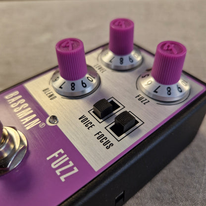 Pre-Owned Fender Bassman Fuzz Pedal Close-up of a Bassman Fuzz bass guitar effects pedal with purple knobs, a purple and silver control panel, and black switches on a gray surface.