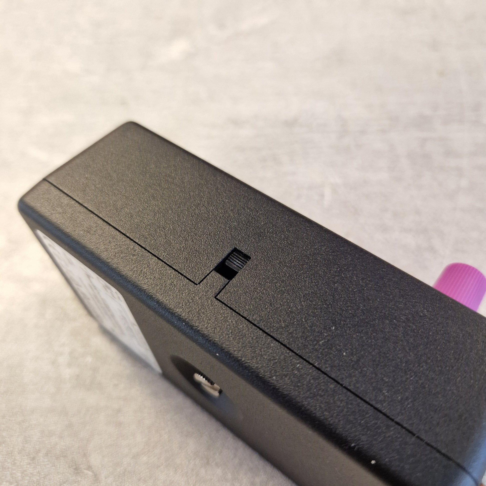 Pre-Owned Fender Bassman Fuzz Pedal Close-up of the top surface of a black rectangular electronic device, showing a textured matte finish, a small sliding switch, a circular port, and a pink connector protruding from the side, set against a light gray textured background.