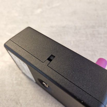 Pre-Owned Fender Bassman Fuzz Pedal Close-up of the top surface of a black rectangular electronic device, showing a textured matte finish, a small sliding switch, a circular port, and a pink connector protruding from the side, set against a light gray textured background.