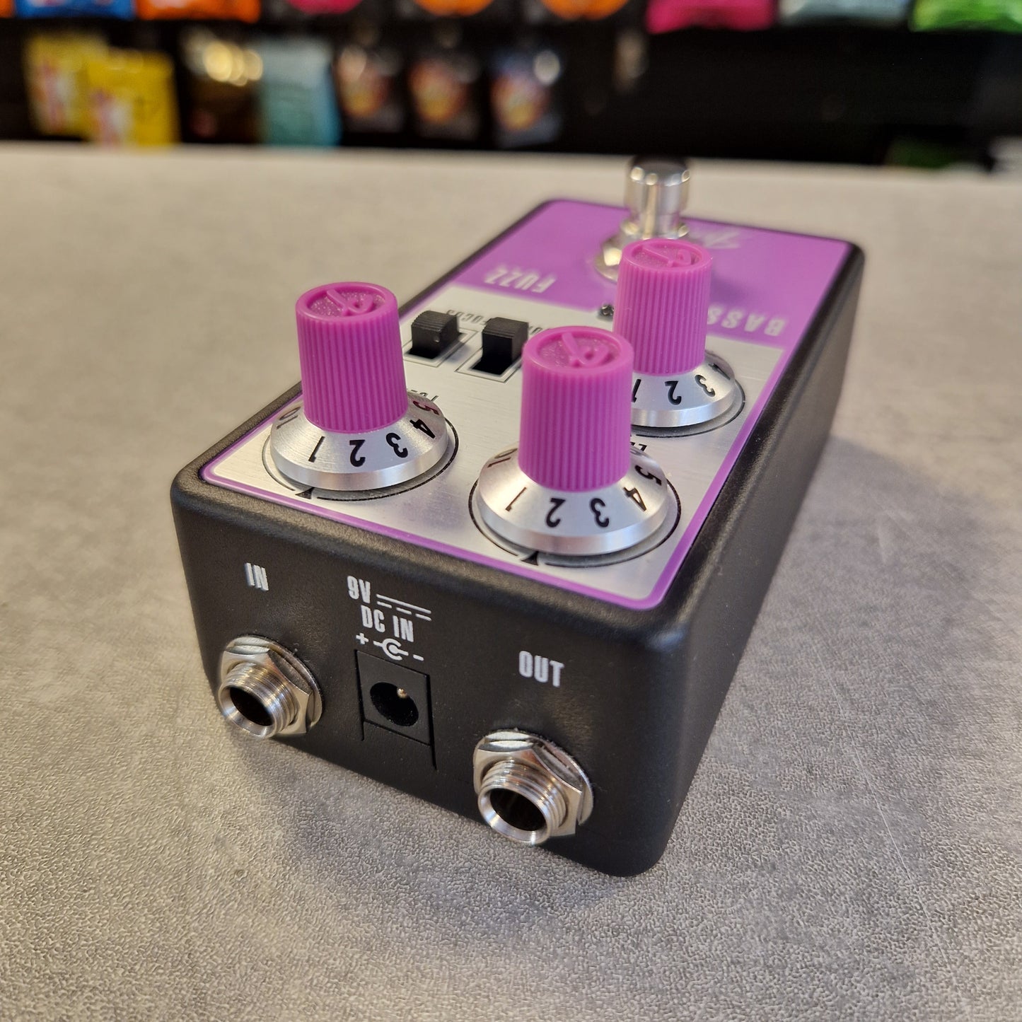 Pre-Owned Fender Bassman Fuzz Pedal A black guitar effects pedal with a purple top plate and three prominent pink-purple rotary knobs, shown on a gray textured surface with a blurred background.