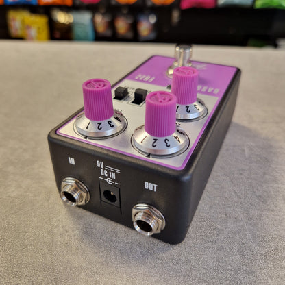 Pre-Owned Fender Bassman Fuzz Pedal A black guitar effects pedal with a purple top plate and three prominent pink-purple rotary knobs, shown on a gray textured surface with a blurred background.