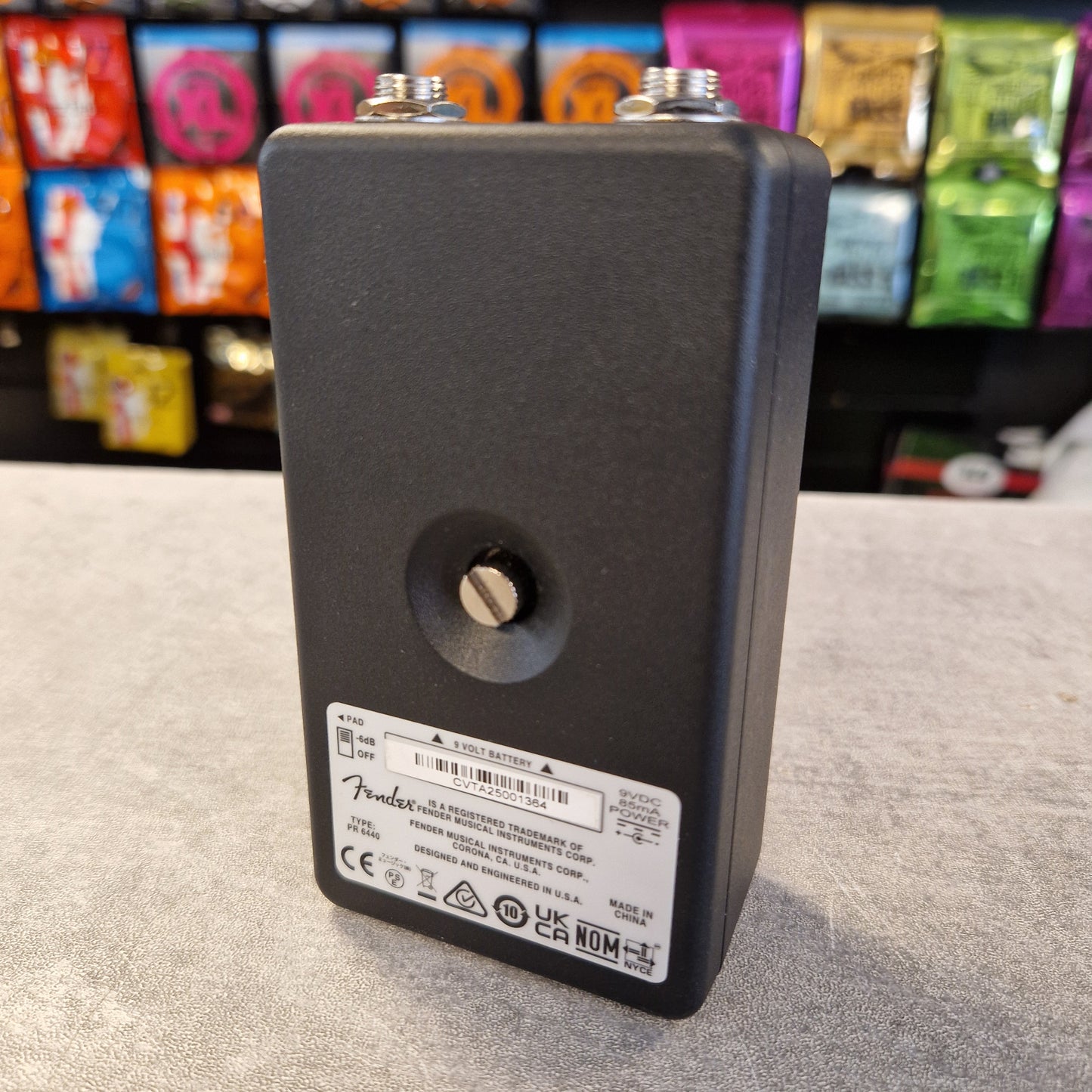 Pre-Owned Fender Bassman Fuzz Pedal A black Fender electric guitar volume control potentiometer, showing the back side with a silver label and metal terminal screws, set on a gray tabletop with a colorful, blurred background.