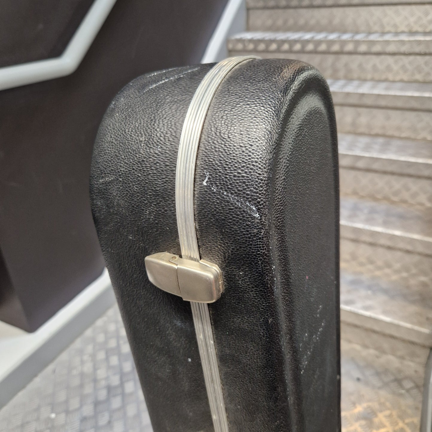 Pre-Owned Hiscox LiteFlite Acoustic Hard Case A close-up of a black textured hard guitar case with a silver metal binding strip and latch, standing on a metal staircase.