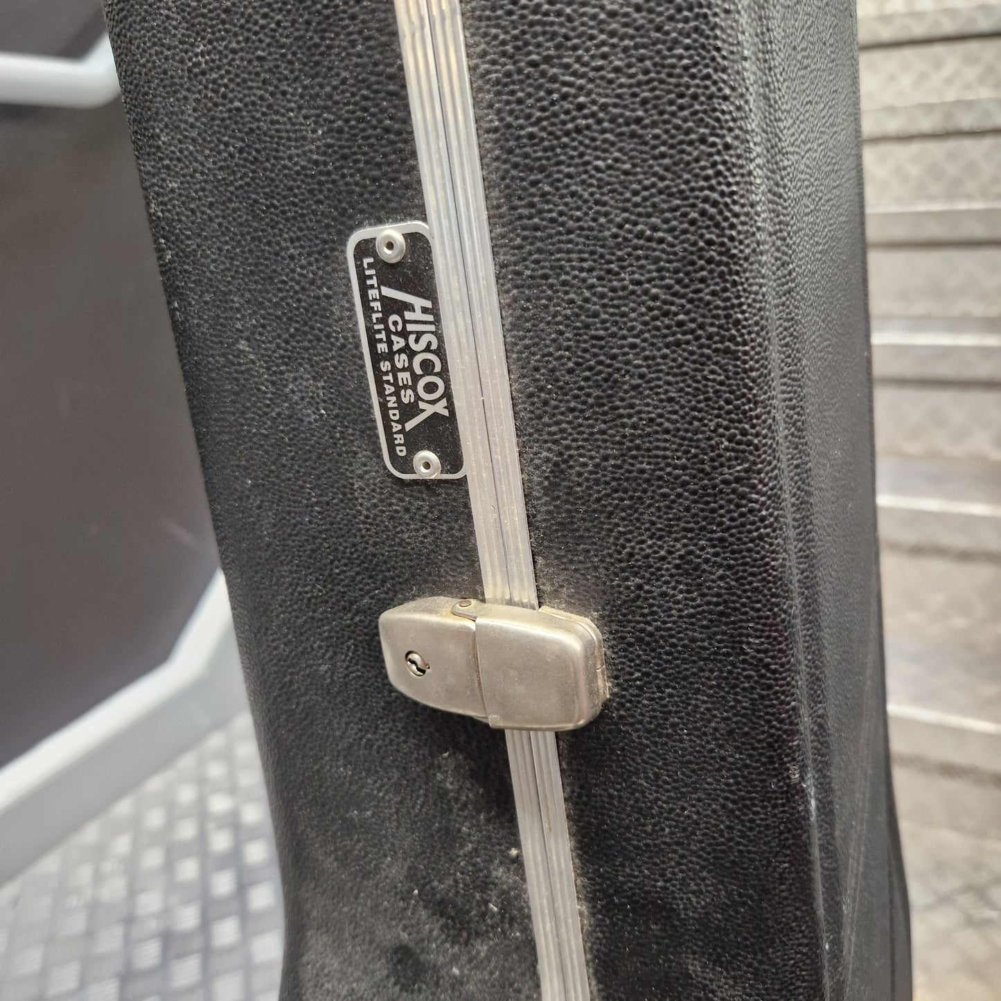 Pre-Owned Hiscox LiteFlite Acoustic Hard Case Close-up of a black textured hard case with a silver metal zipper track, silver latch, and a visible Misco brand plate.