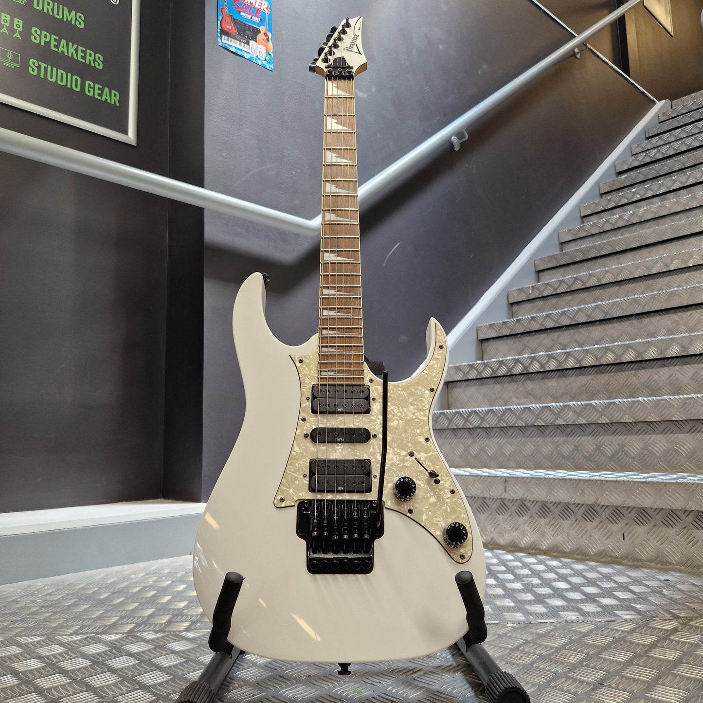 Pre-Owned Ibanez RG350DX Electric - White A white electric guitar with a black bridge and pickups, displayed on a stand in front of a staircase.