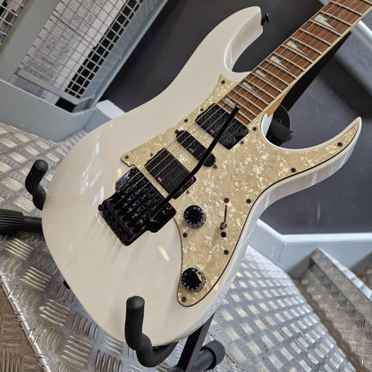 Pre-Owned Ibanez RG350DX Electric - White A white electric guitar with a pearlescent cream pickguard and black hardware, resting on a metal stand in what appears to be a showroom or workshop.