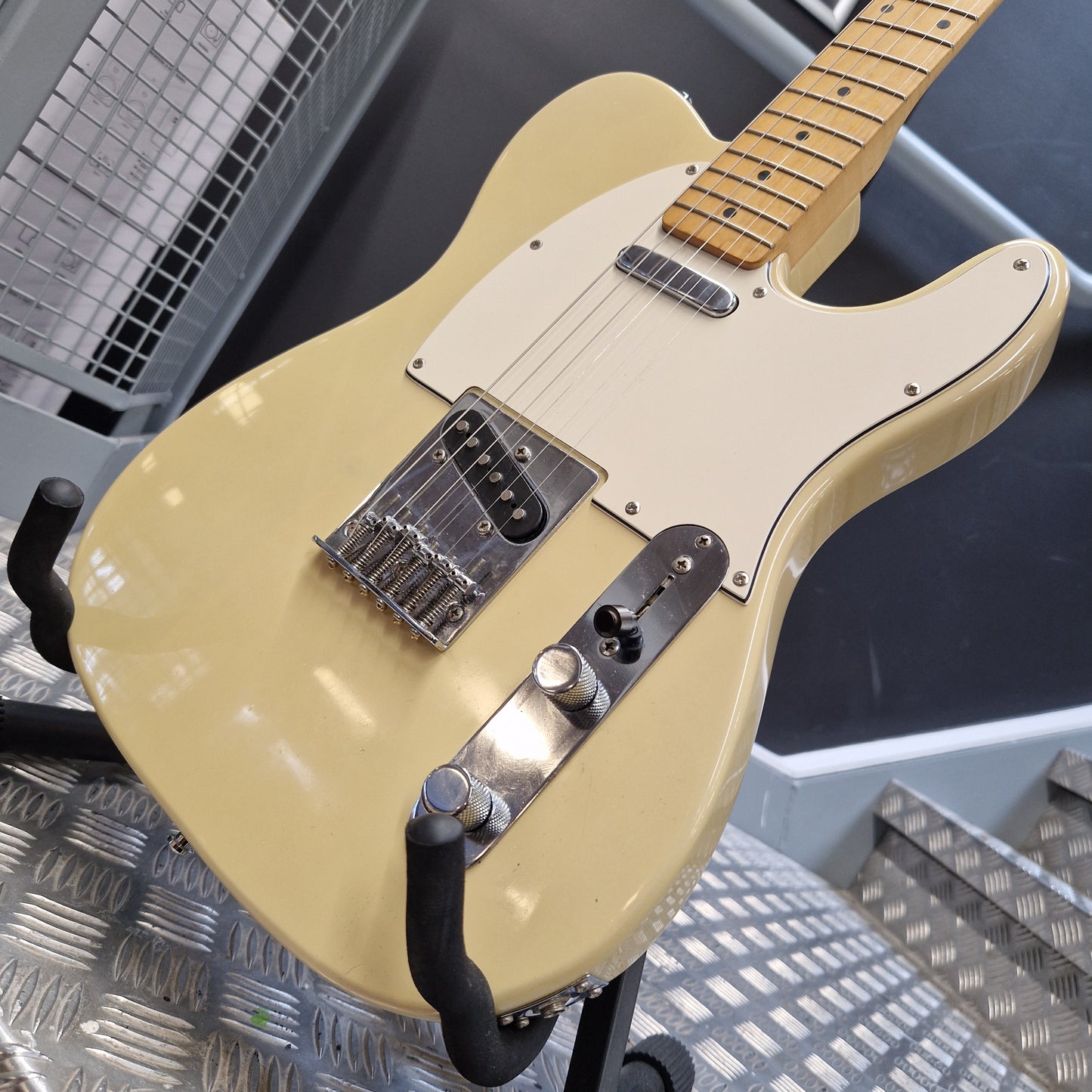 Pre-Owned Squier MIK 1996 Telecaster Electric - Blonde A cream-colored electric guitar with a maple neck, resting on a black stand over a diamond-plate metal floor.