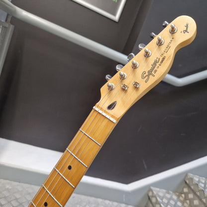 Pre-Owned Squier MIK 1996 Telecaster Electric - Blonde Close-up of a Fender Stratocaster electric guitar headstock and neck, showing the maple wood headstock, six silver tuning pegs, and the Fender logo, set against a dark gray wall and metal steps.