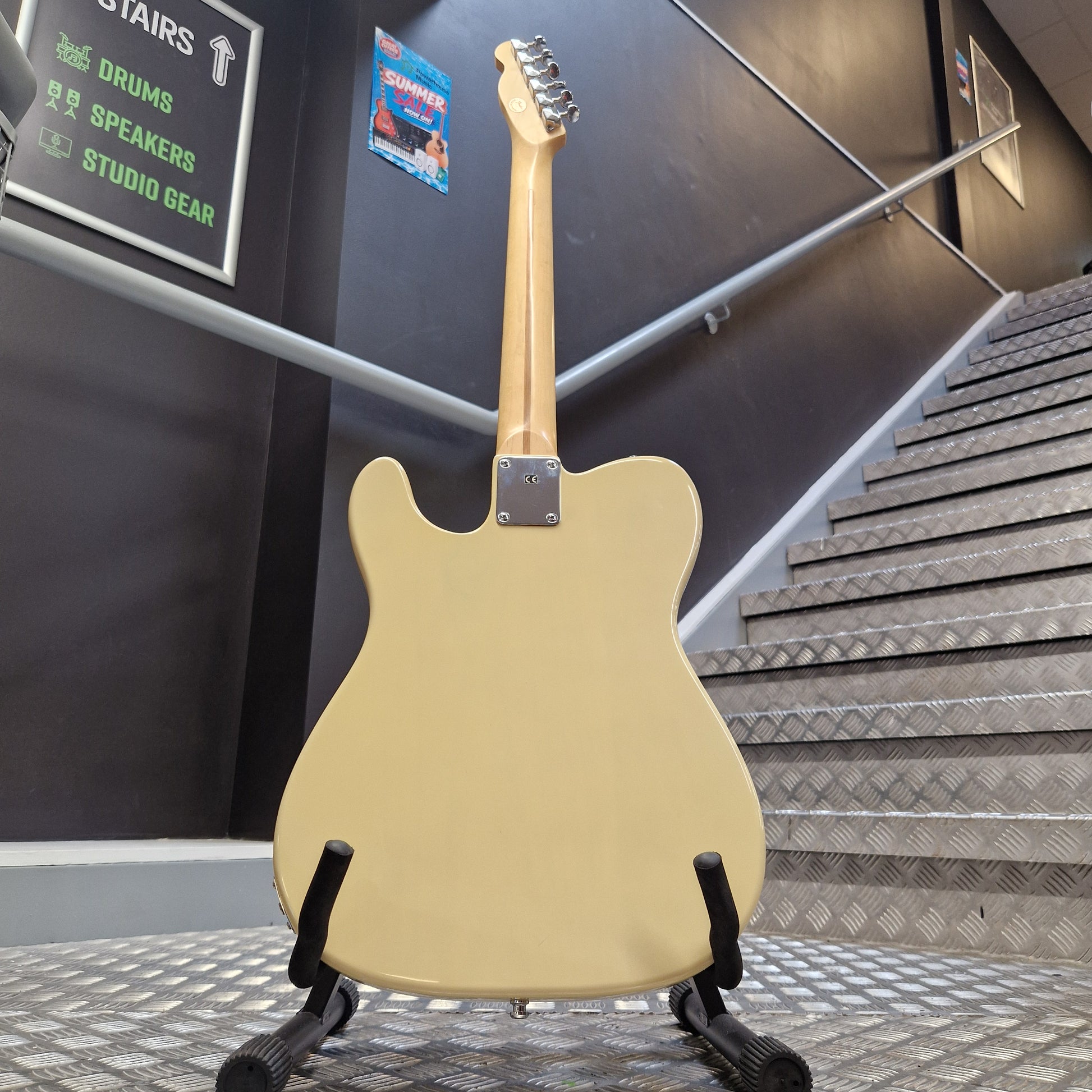Pre-Owned Squier MIK 1996 Telecaster Electric - Blonde A single-cutaway electric guitar with a cream-colored body and a natural wood neck, displayed on a black stand in an indoor setting with a staircase in the background.
