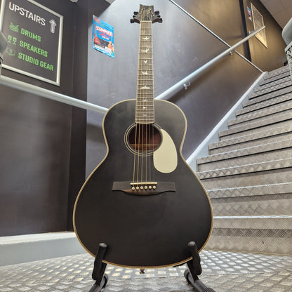 Pre-Owned PRS P20 Tonare Parlor Electro-Acoustic - Black A black acoustic guitar with gold-toned hardware and a white pickguard, displayed on a stand in front of a staircase.