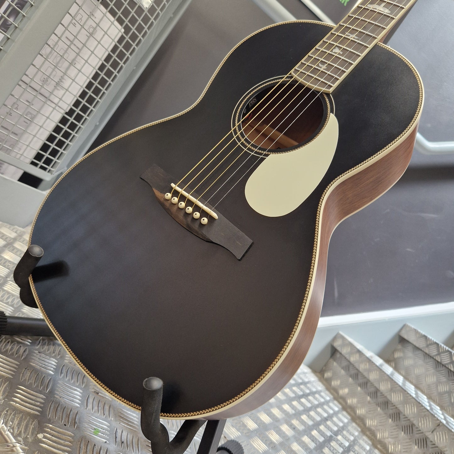 Pre-Owned PRS P20 Tonare Parlor Electro-Acoustic - Black A close-up of an acoustic guitar body on a stand, featuring a dark brown to black varnished finish, a cream pickguard, and gold-toned hardware, photographed in what appears to be a showroom or studio setting.