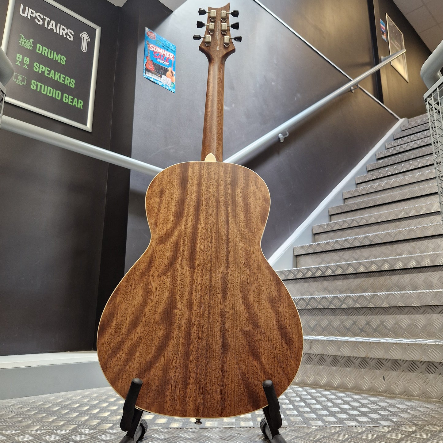 Pre-Owned PRS P20 Tonare Parlor Electro-Acoustic - Black The back of a wooden acoustic guitar with a glossy brown finish and visible grain, displayed on a stand in an indoor stairwell environment.