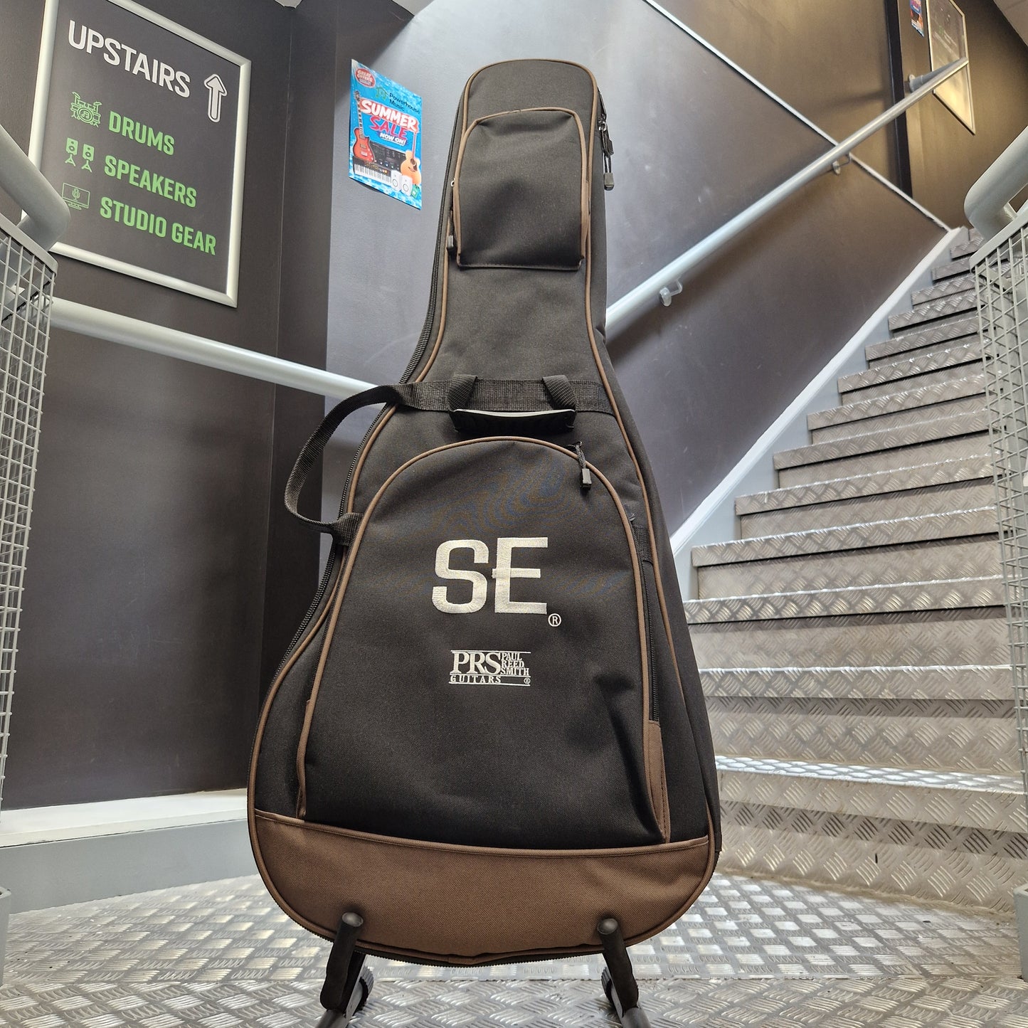 Pre-Owned PRS P20 Tonare Parlor Electro-Acoustic - Black A PRS guitar gig bag with a black and brown design, featuring a large white 'SE' logo and PRS branding, standing on a metal staircase landing.