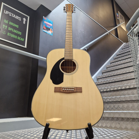 Pre-Owned Fender CD-60S Dreadnought Acoustic Left-Handed - Natural A natural wood acoustic guitar with a black pickguard is displayed on a stand in front of a staircase and dark gray wall.