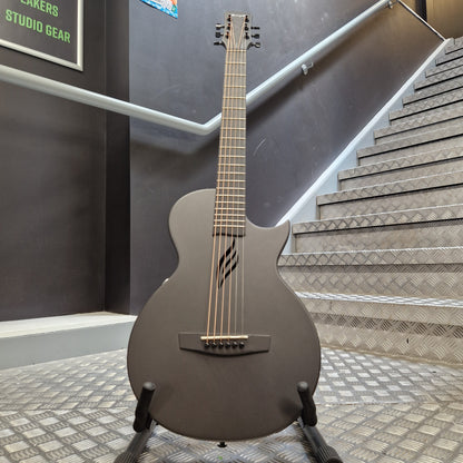 Pre-Owned Enya Nova Go SP1 Carbon Fibre Electro-Acoustic - Matte Black A matte black cutaway electric guitar on a stand, positioned in front of a staircase and dark gray wall.