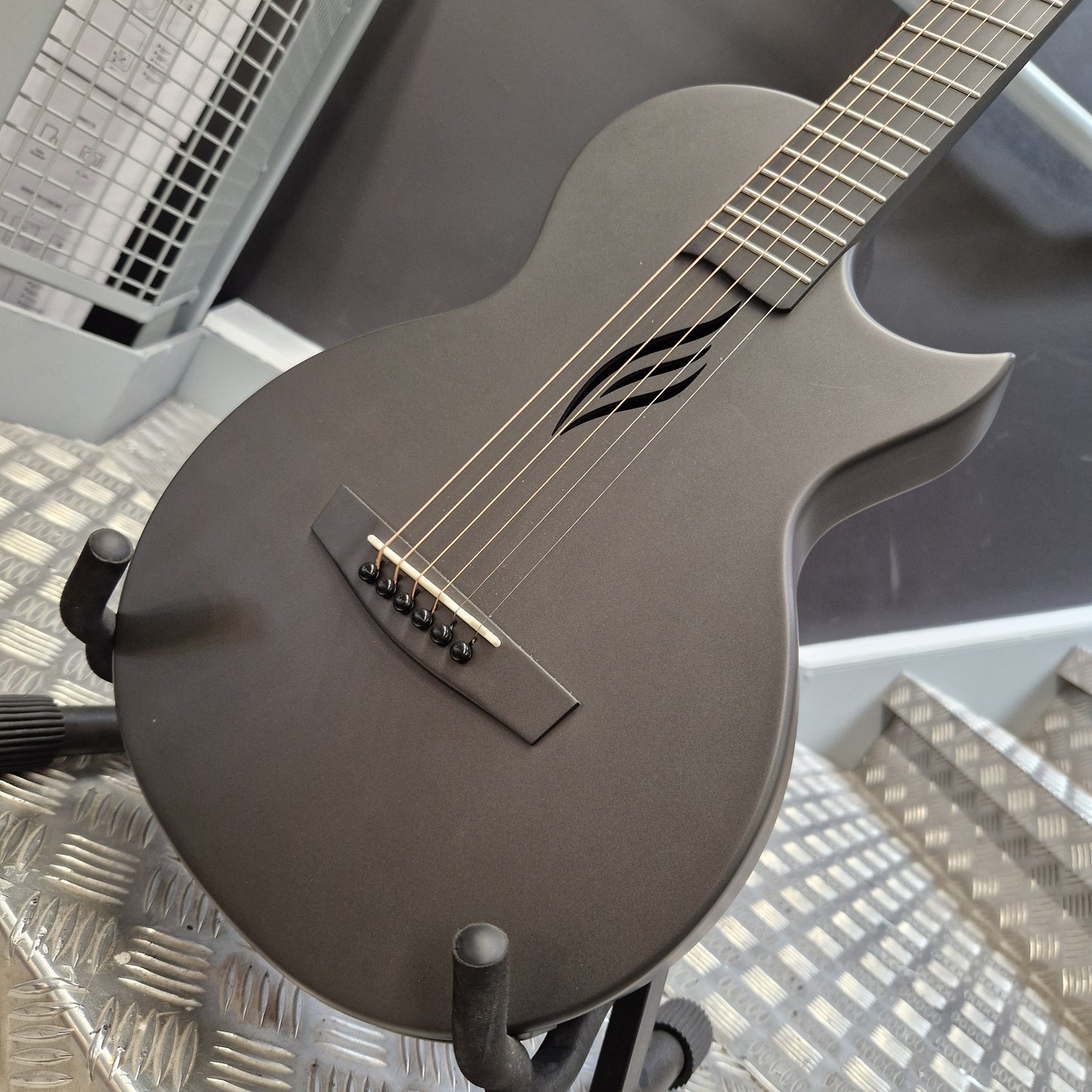 Pre-Owned Enya Nova Go SP1 Carbon Fibre Electro-Acoustic - Matte Black Matte black electric guitar with a distinctive cutaway body and decorative soundhole, resting on a black stand on a textured metal surface.