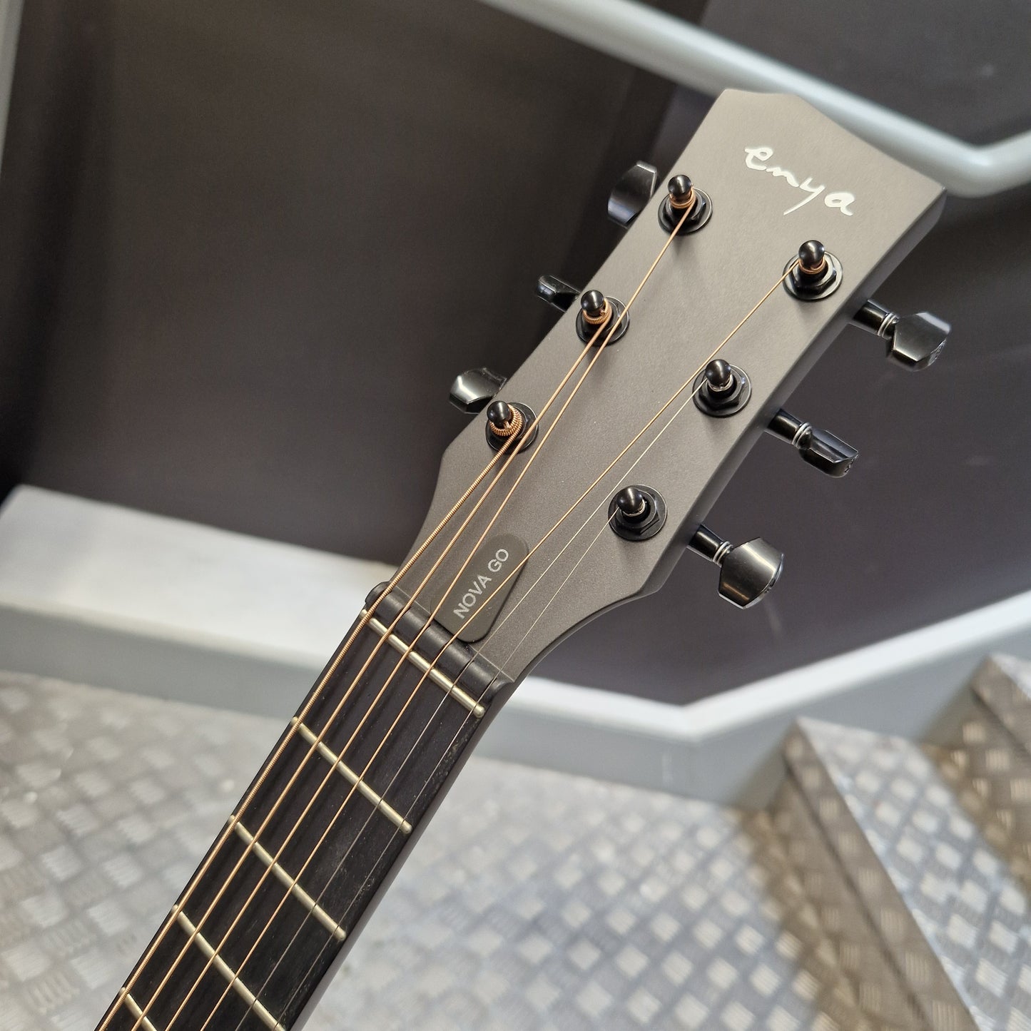 Pre-Owned Enya Nova Go SP1 Carbon Fibre Electro-Acoustic - Matte Black Close-up of the headstock of an Enya Nova Co acoustic guitar, showing the brand name 'enya' in white script and 'NOVA CO' on the side, with black tuning pegs and strings against a dark and light gray background.