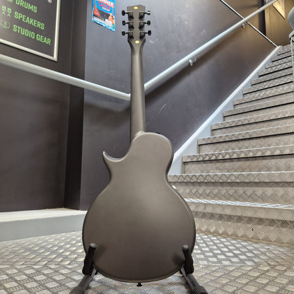 Pre-Owned Enya Nova Go SP1 Carbon Fibre Electro-Acoustic - Matte Black A matte dark gray electric guitar shown from the back, resting on a black stand in front of an indoor staircase with metal steps and a handrail.