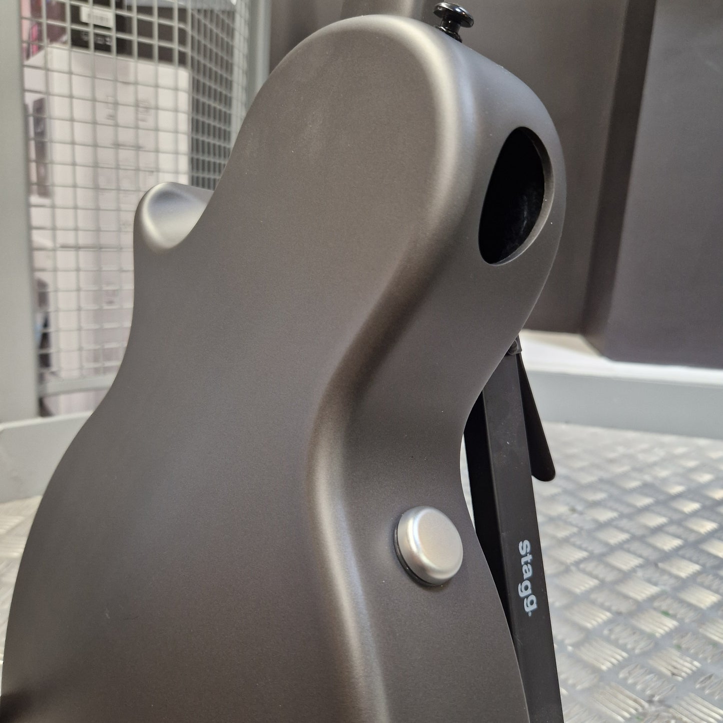 Pre-Owned Enya Nova Go SP1 Carbon Fibre Electro-Acoustic - Matte Black Close-up of a matte dark gray electric guitar body showing the lower bout, a silver control knob, and part of a black pickguard with white 'stage' branding, set against a workshop or studio background.