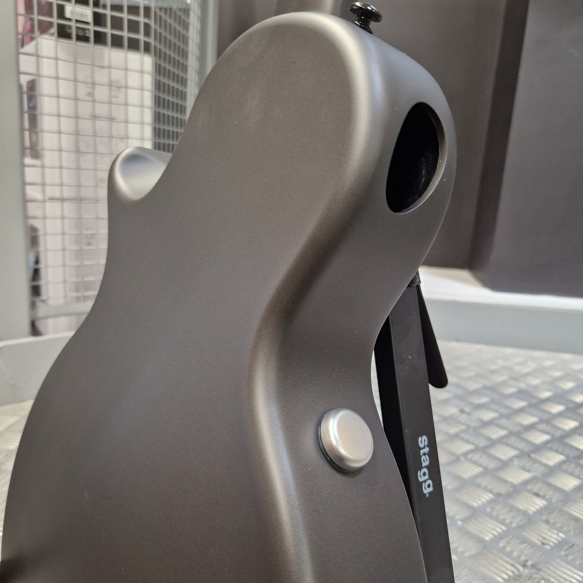 Pre-Owned Enya Nova Go SP1 Carbon Fibre Electro-Acoustic - Matte Black Close-up of a matte dark gray electric guitar body showing the lower bout, a silver control knob, and part of a black pickguard with white 'stage' branding, set against a workshop or studio background.