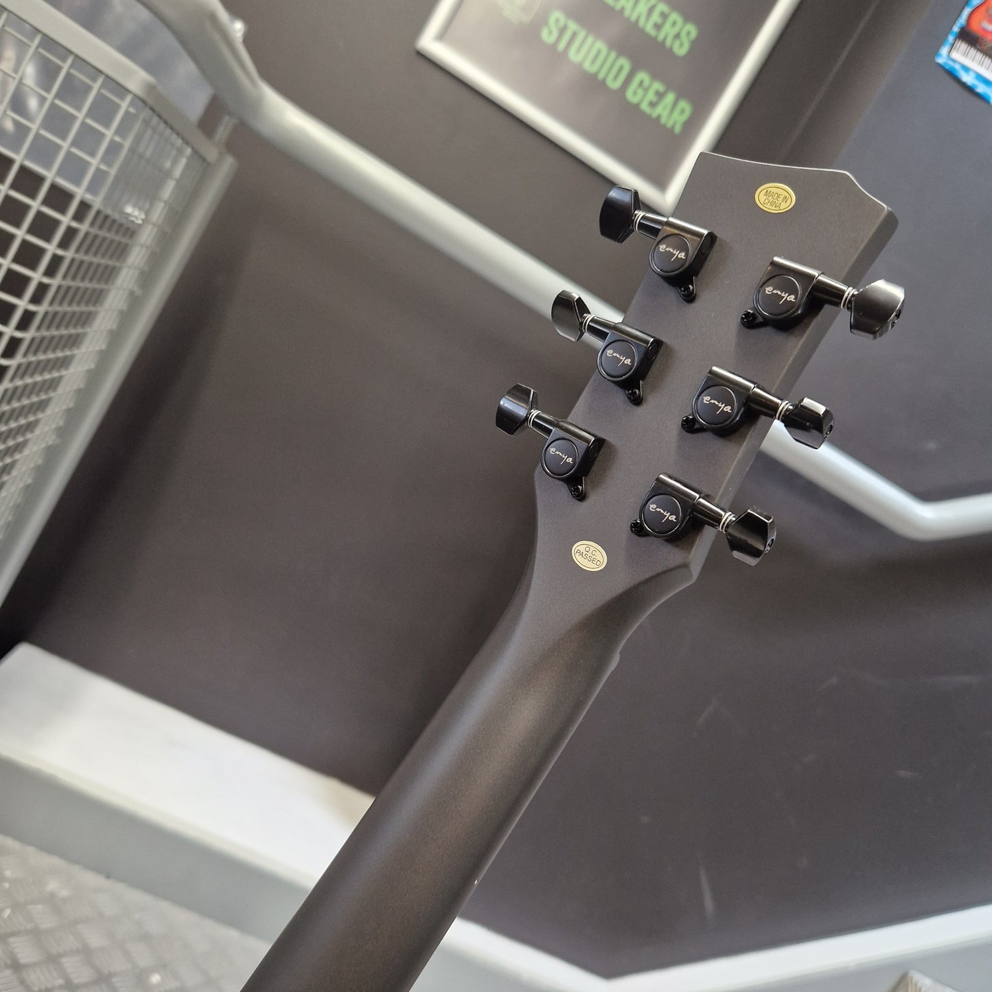 Pre-Owned Enya Nova Go SP1 Carbon Fibre Electro-Acoustic - Matte Black Close-up of the headstock of a dark gray or black acoustic guitar, showing six tuning pegs and two gold oval stickers, set against a dark gray studio background with a sign that reads 'STUDIO GEAR'.