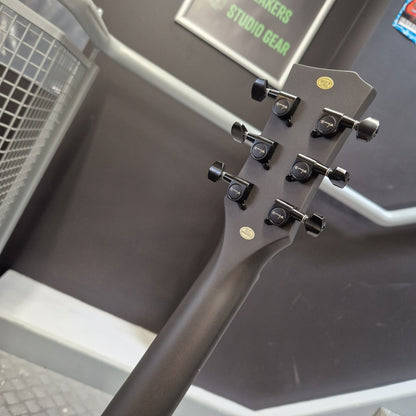 Pre-Owned Enya Nova Go SP1 Carbon Fibre Electro-Acoustic - Matte Black Close-up of the headstock of a dark gray or black acoustic guitar, showing six tuning pegs and two gold oval stickers, set against a dark gray studio background with a sign that reads 'STUDIO GEAR'.