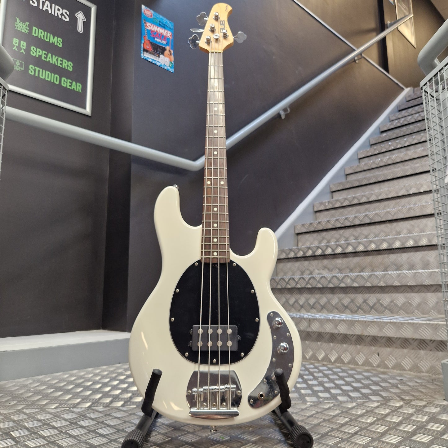 Pre-Owned Music Man Sterling Sub Series Ray 4 Bass - White An off-white electric bass guitar with a black pickguard and chrome hardware, standing on a black stand in front of a staircase.