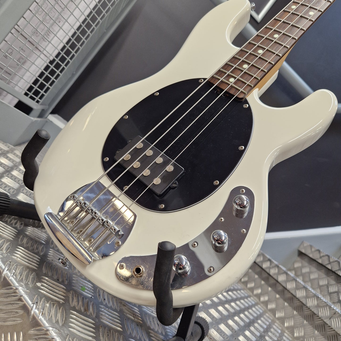 Pre-Owned Music Man Sterling Sub Series Ray 4 Bass - White A close-up of a white electric bass guitar with a black pickguard, chrome hardware, and a dark wood fretboard, resting on a metal stand.