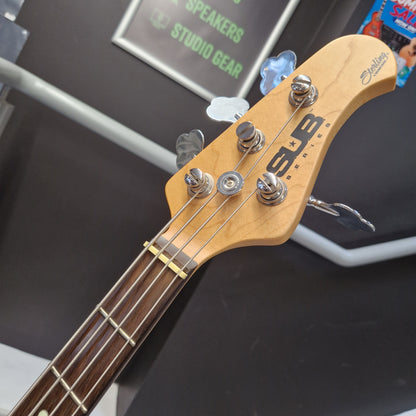 Pre-Owned Music Man Sterling Sub Series Ray 4 Bass - White Close-up of a four-string bass guitar headstock with a natural wood finish, chrome tuning pegs, and a dark fretboard, photographed in a store or showroom setting.