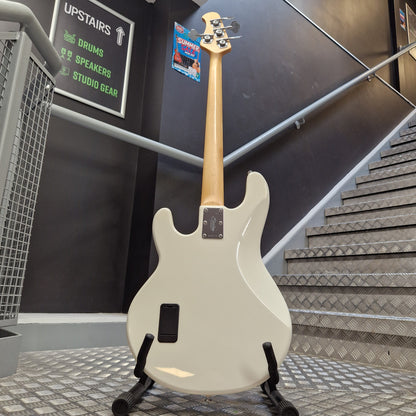 Pre-Owned Music Man Sterling Sub Series Ray 4 Bass - White A white electric bass guitar with a natural wood neck stands on a black stand in a shop or studio setting with stairs and signage in the background.