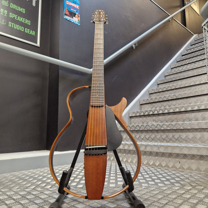 Pre-Owned Yamaha SLG200S Silent Guitar Electro-Acoustic - Natural A unique contoured electric guitar with a warm brown wood finish and black hardware, displayed on a stand in front of a staircase.
