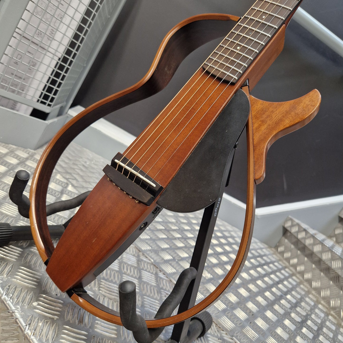 Pre-Owned Yamaha SLG200S Silent Guitar Electro-Acoustic - Natural A wooden six-string acoustic guitar with a distinctive curved body and black headstock, displayed on a black stand on a diamond plate floor.
