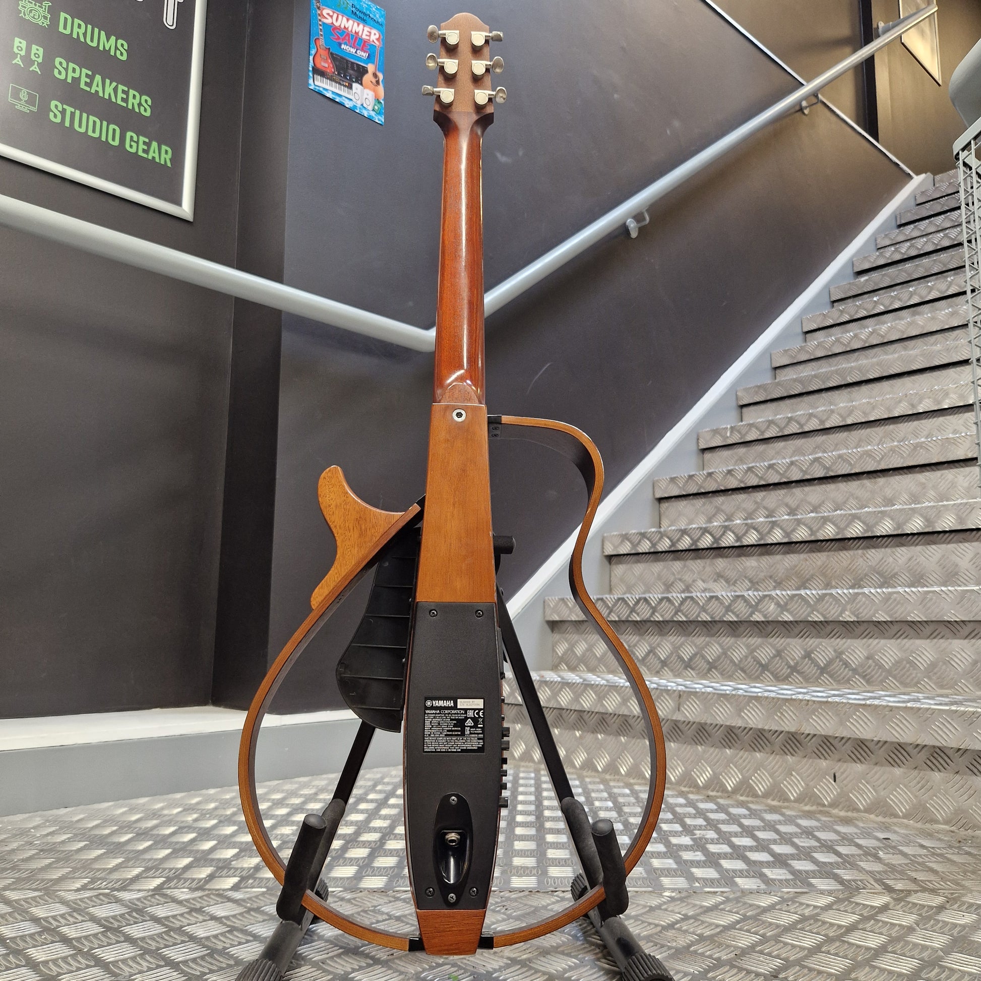 Pre-Owned Yamaha SLG200S Silent Guitar Electro-Acoustic - Natural A Yamaha electric guitar stand holding a wooden acoustic-electric style guitar, photographed indoors against a staircase and dark wall with signage in the background.