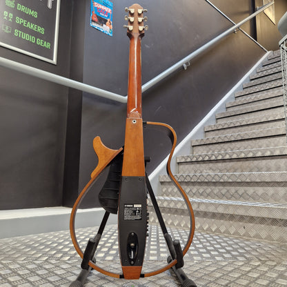Pre-Owned Yamaha SLG200S Silent Guitar Electro-Acoustic - Natural A Yamaha electric guitar stand holding a wooden acoustic-electric style guitar, photographed indoors against a staircase and dark wall with signage in the background.