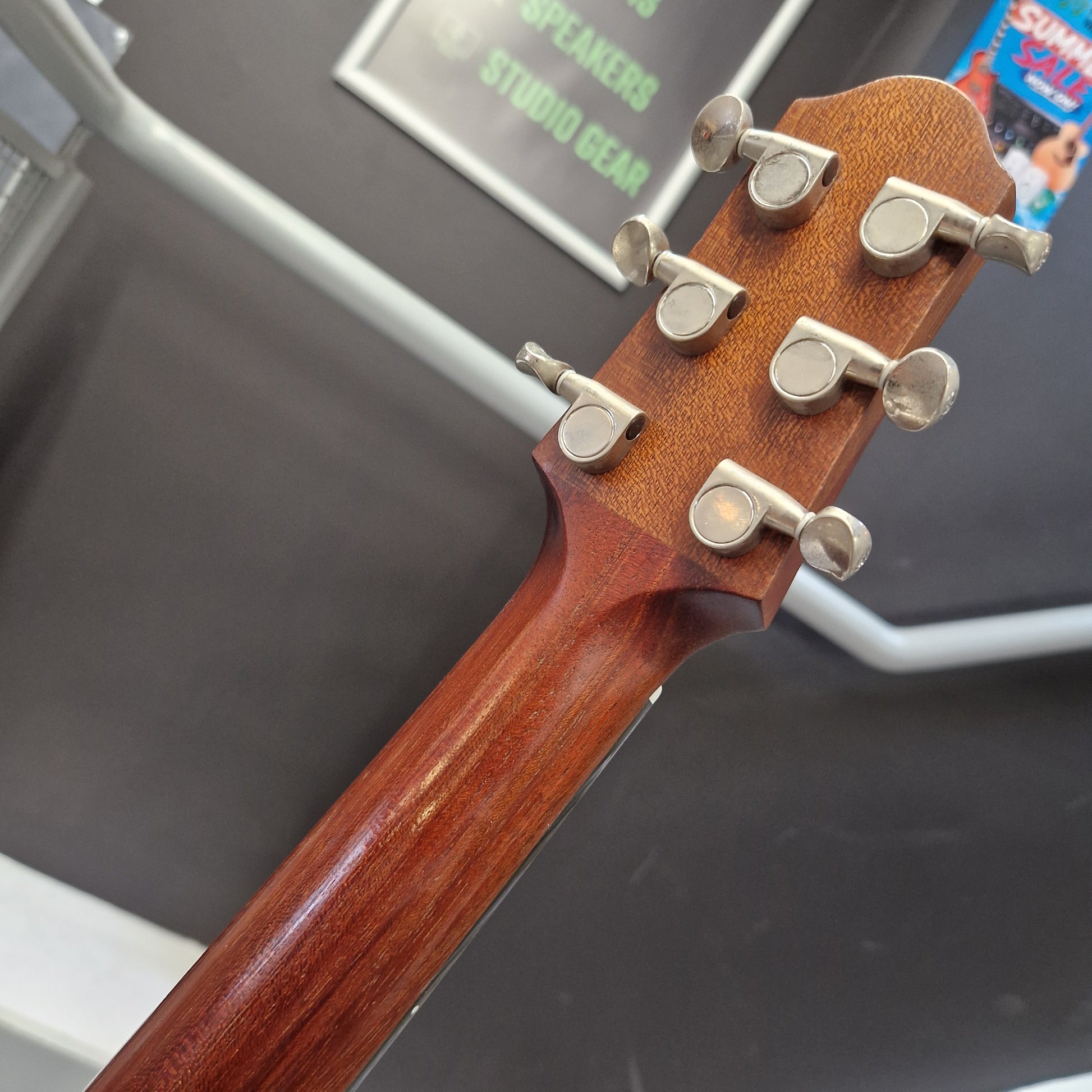 Pre-Owned Yamaha SLG200S Silent Guitar Electro-Acoustic - Natural Close-up of the headstock of an acoustic guitar, showing six silver tuning pegs and a warm reddish-brown wooden finish, set against a dark gray background with store signage.