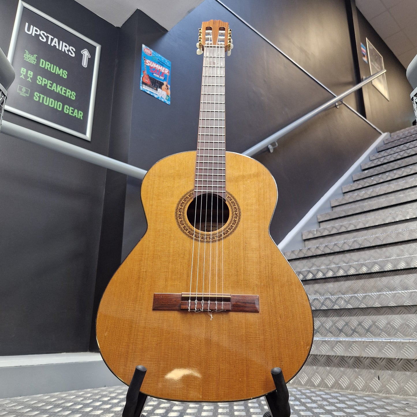 Pre-Owned Admira Solista Classical - Natural A classical acoustic guitar with a natural wood finish is displayed on a stand in a stairwell. The guitar features a warm honey-toned wooden body, dark brown neck and headstock, and a decorative rosette around the sound hole.