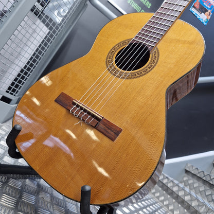 Pre-Owned Admira Solista Classical - Natural A close-up of a classical guitar body made from natural wood, showing the grain, sound hole, bridge, and part of the neck, displayed on a stand in what appears to be a workshop or studio.