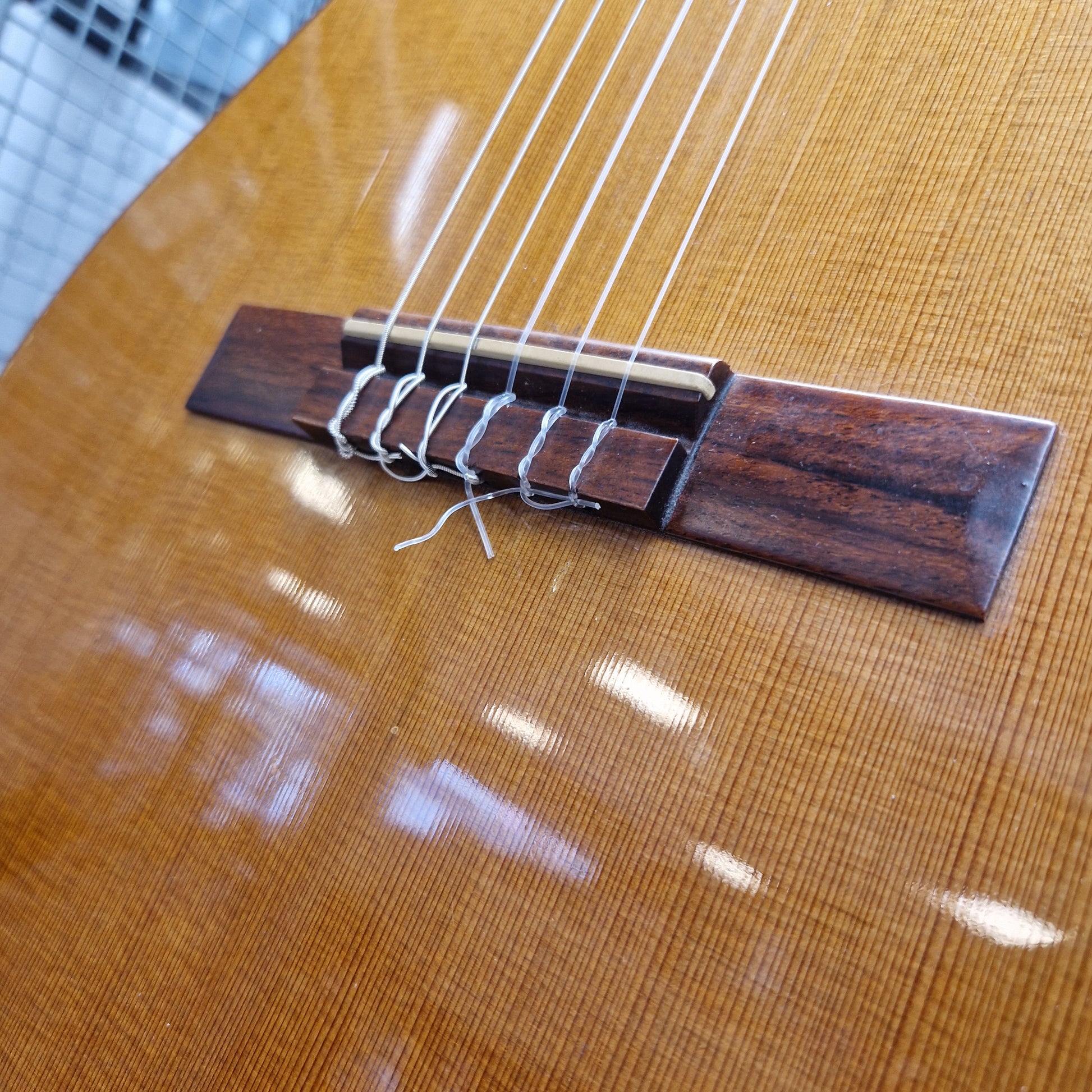 Pre-Owned Admira Solista Classical - Natural Close-up of a guitar's bridge and strings, showing a dark brown wooden bridge with silver metal string trees and silver strings against a glossy golden-brown wood body.
