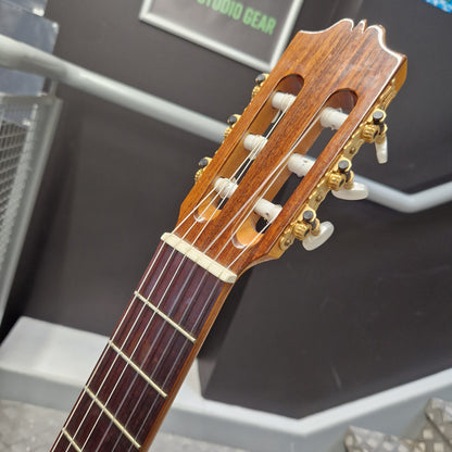 Pre-Owned Admira Solista Classical - Natural Close-up of a classical guitar headstock and upper neck, showing six tuning pegs, four strings, and a warm brown wood finish, photographed indoors against a dark gray wall.