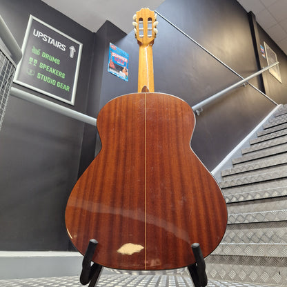 Pre-Owned Admira Solista Classical - Natural A classical guitar shown from the back, resting on a black display stand in a stairwell. The back panel has a warm reddish-brown wood finish with visible grain and a small chip near the lower center.