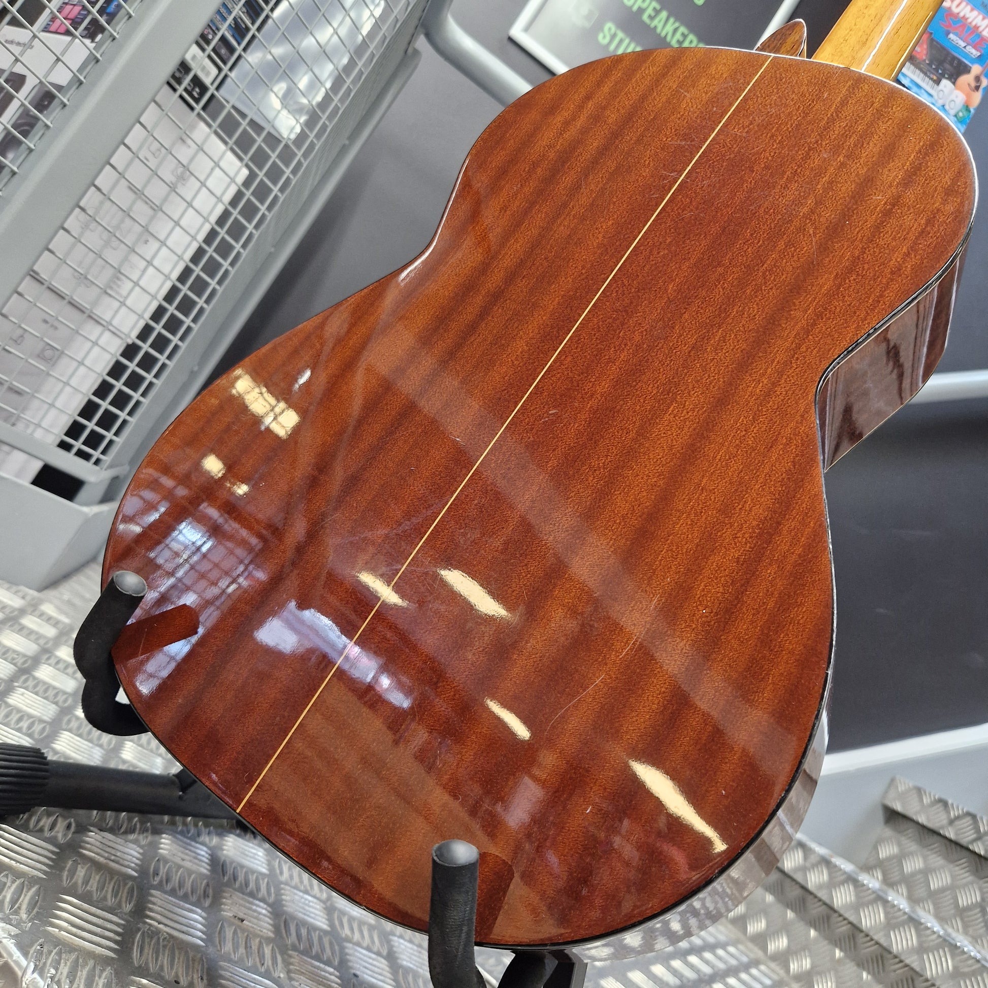 Pre-Owned Admira Solista Classical - Natural Close-up of the glossy back of an acoustic guitar showing a rich reddish-brown wood grain finish, displayed on a stand in a store setting.