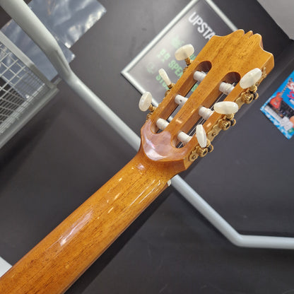 Pre-Owned Admira Solista Classical - Natural Close-up of the headstock and neck of a classical guitar with a glossy warm brown wood finish and white tuning pegs, photographed indoors against a dark display background.