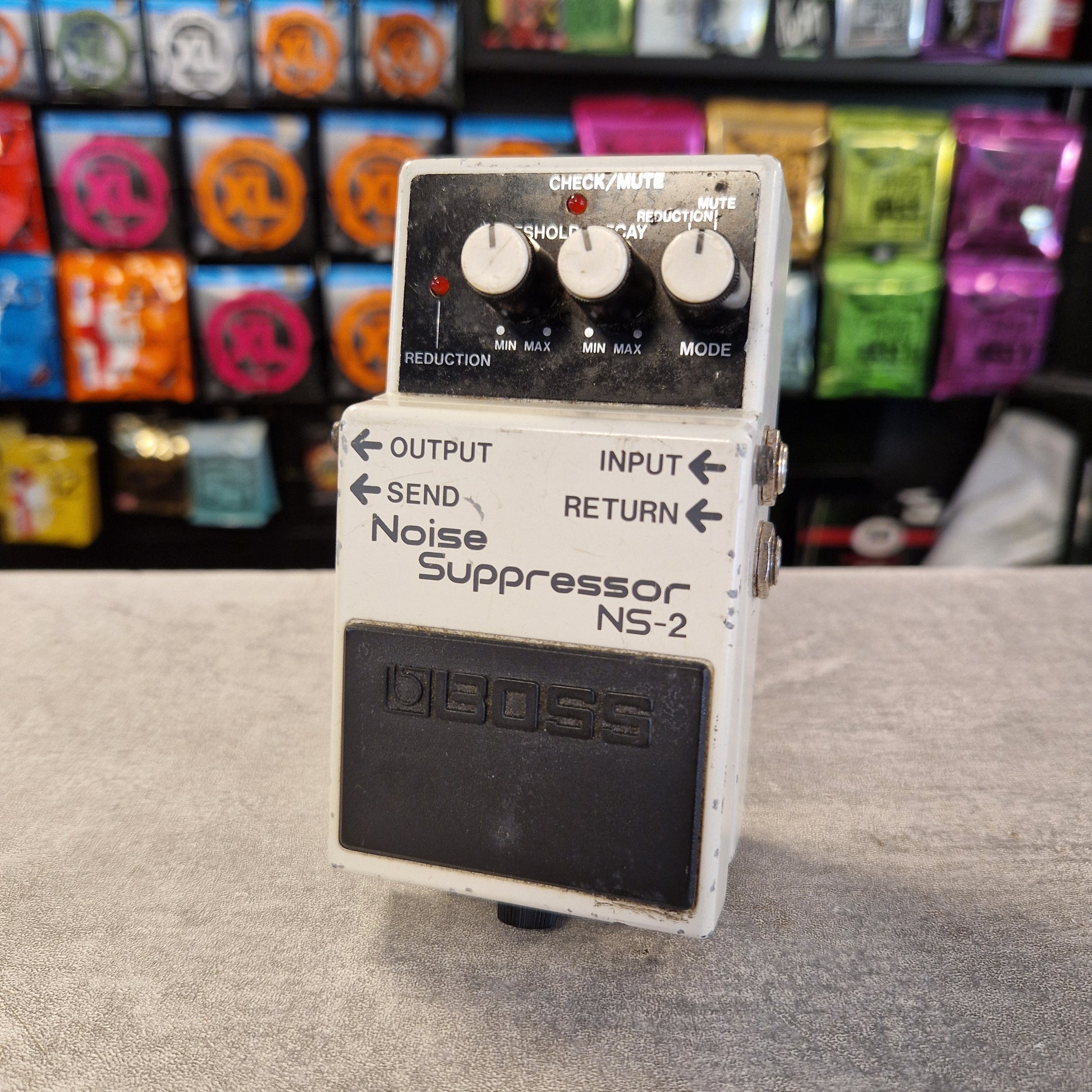 Pre-Owned Boss NS-2 Noise Suppressor Pedal A vintage Boss NS-2 Noise Suppressor guitar pedal with a white metal casing, black footplate, and three white control knobs, set against a blurred background of colorful guitar parts.