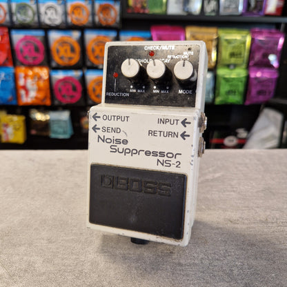 Pre-Owned Boss NS-2 Noise Suppressor Pedal A vintage Boss NS-2 Noise Suppressor guitar pedal with a white metal casing, black footplate, and three white control knobs, set against a blurred background of colorful guitar parts.