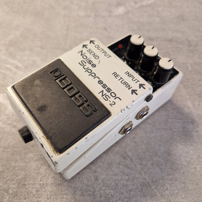 Pre-Owned Boss NS-2 Noise Suppressor Pedal A vintage BOSS Noise Suppressor NS-2 guitar effects pedal with a white body, black footswitch, and three black knobs with white tops, resting on a gray textured surface.
