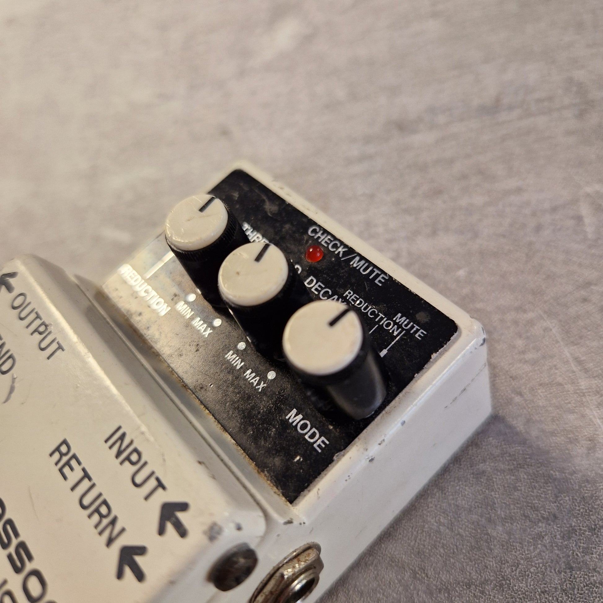 Pre-Owned Boss NS-2 Noise Suppressor Pedal A close-up of a white guitar effects pedal with black knobs and a black control panel, placed on a gray textured surface. Part of another pedal is visible at the left edge.