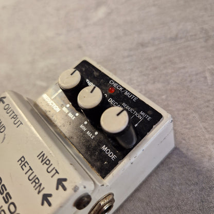 Pre-Owned Boss NS-2 Noise Suppressor Pedal A close-up of a white guitar effects pedal with black knobs and a black control panel, placed on a gray textured surface. Part of another pedal is visible at the left edge.
